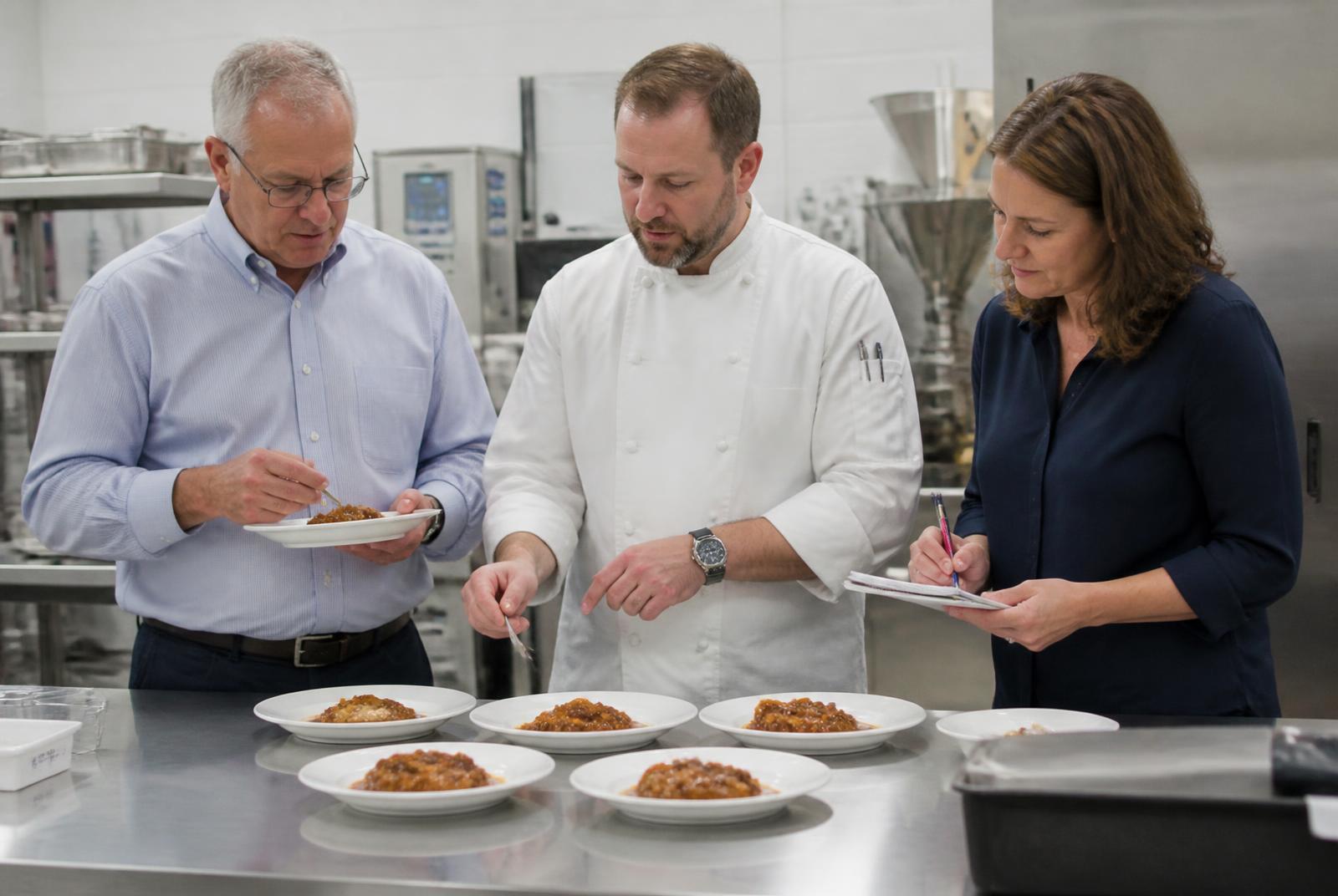 An R&D chef tasting and comparing food samples at a development bench with two food manufacturing customer representatives taking notes
